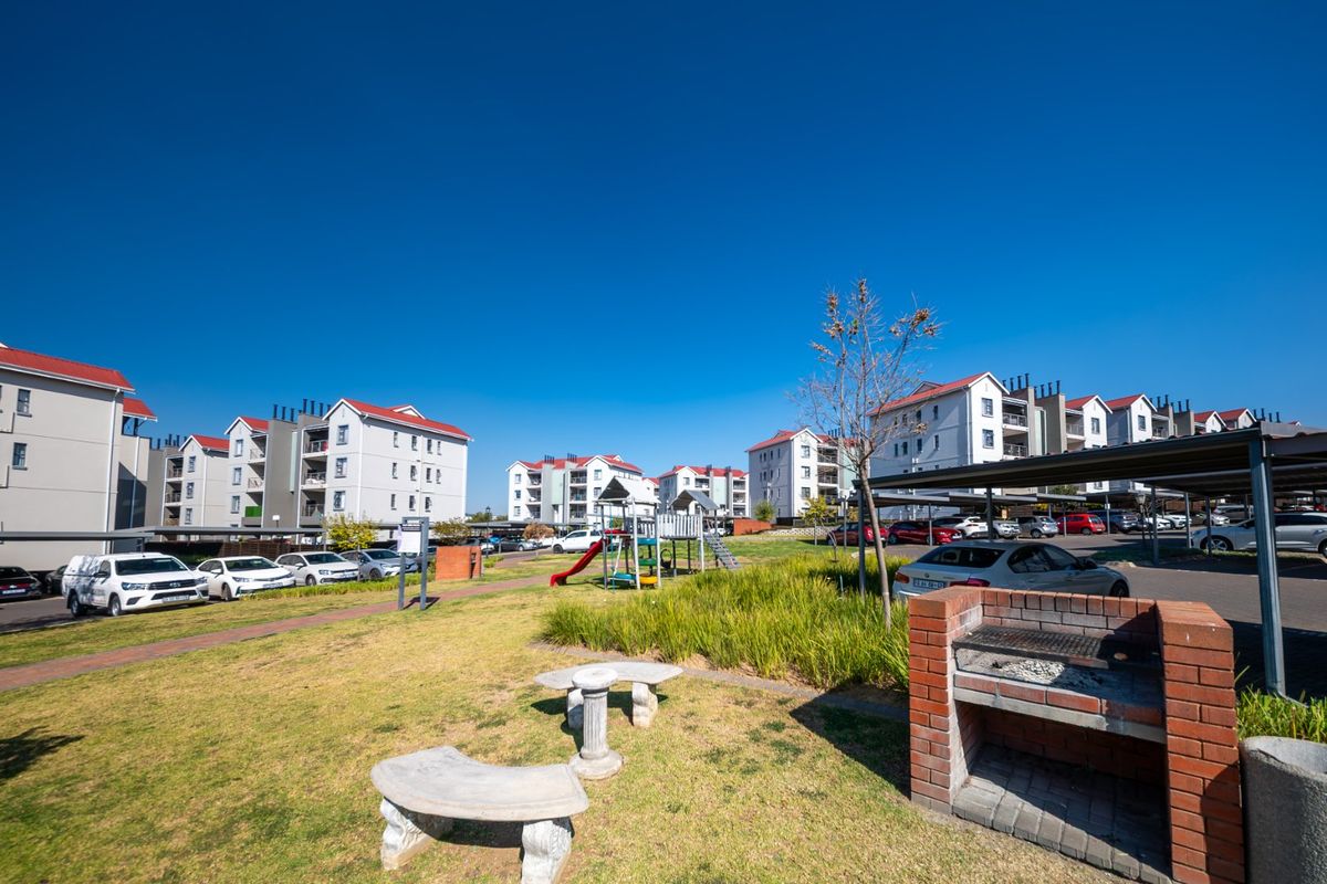 Communal outdoor area featuring a brick built-in braai, concrete seating, and a children's playground set within an apartment complex.