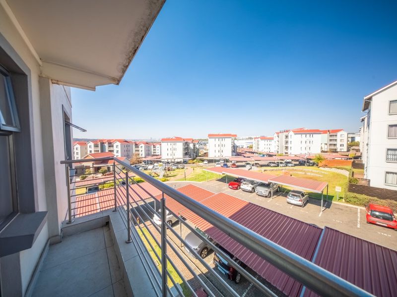 A private balcony featuring tiled flooring and a stainless steel balustrade, offering an elevated view over the apartment complex parking area.