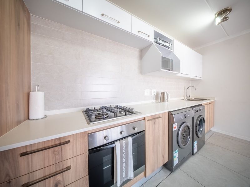 Modern kitchen featuring light wood cabinetry, stone countertops, a gas hob, built-in oven, and integrated laundry appliance space with a tiled backsplash.