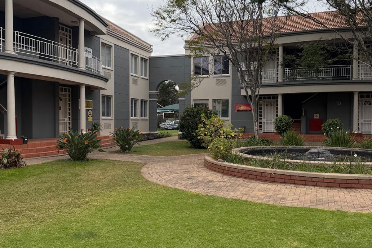 A well-maintained commercial office complex courtyard featuring landscaped lawns, a central water feature, paved walkways, and multi-story buildings with balconies.