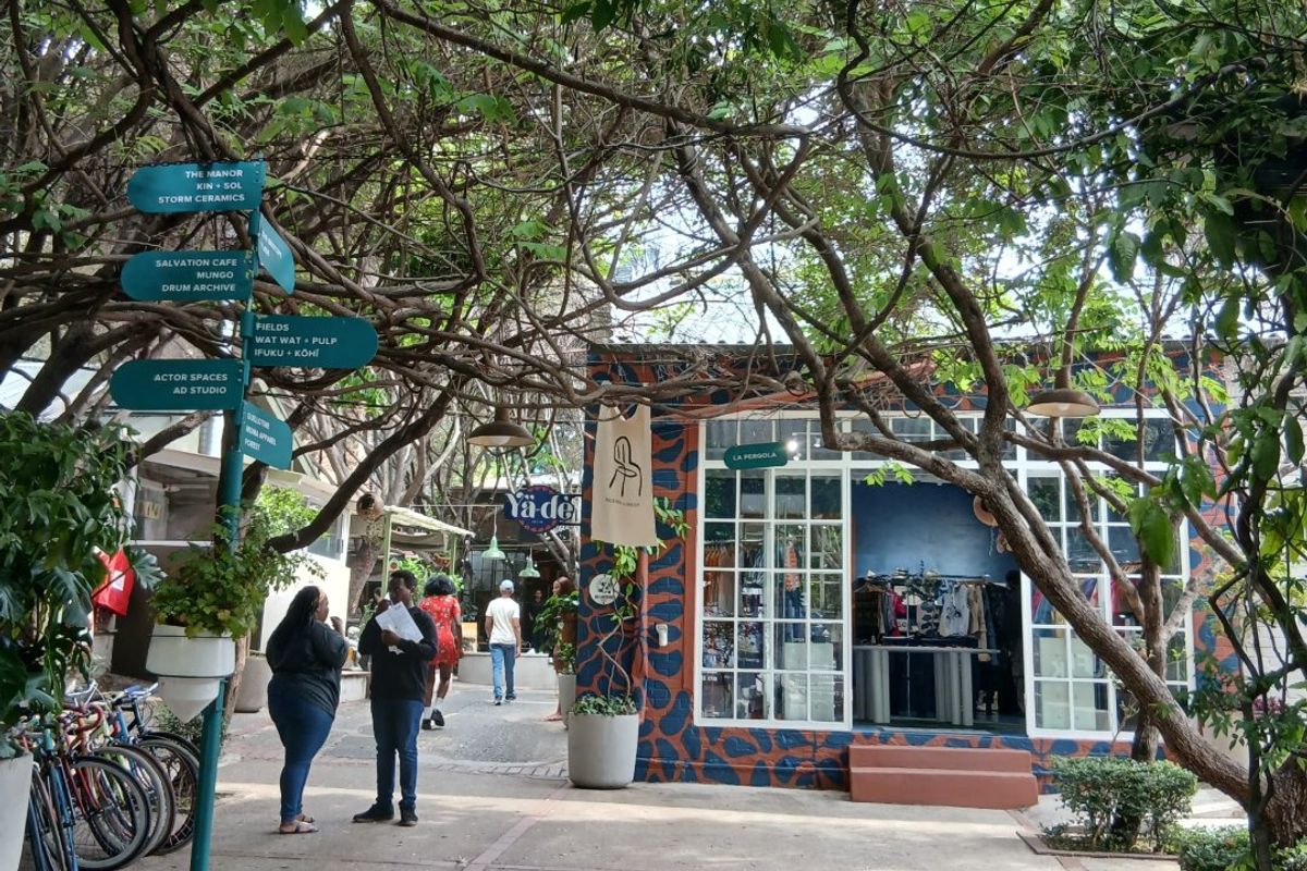A vibrant outdoor courtyard area featuring mature trees, paved walkways, and commercial storefronts with large glass windows.