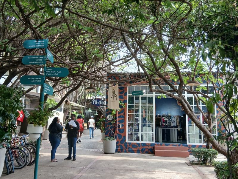 A vibrant outdoor courtyard area featuring mature trees, paved walkways, and commercial storefronts with large glass windows.