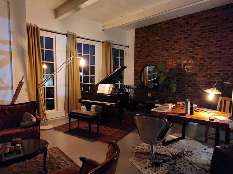 Spacious living area featuring an exposed brick feature wall, high ceilings with exposed beams, and large windows providing ample natural light.