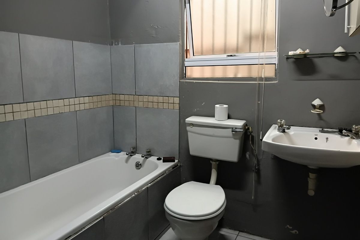 A functional bathroom featuring a bathtub, pedestal basin, and toilet, with grey wall finishes and tiled splashbacks requiring cosmetic updates.