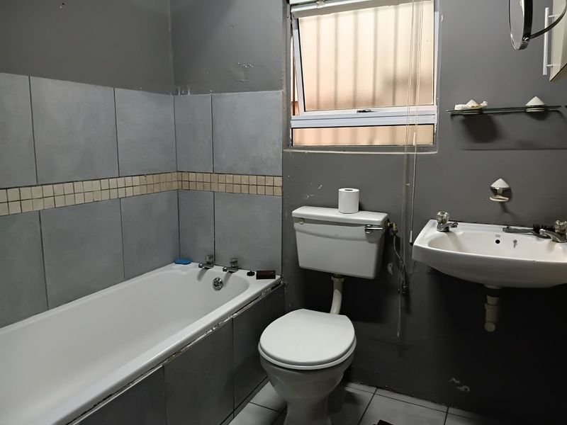 A functional bathroom featuring a bathtub, pedestal basin, and toilet, with grey wall finishes and tiled splashbacks requiring cosmetic updates.