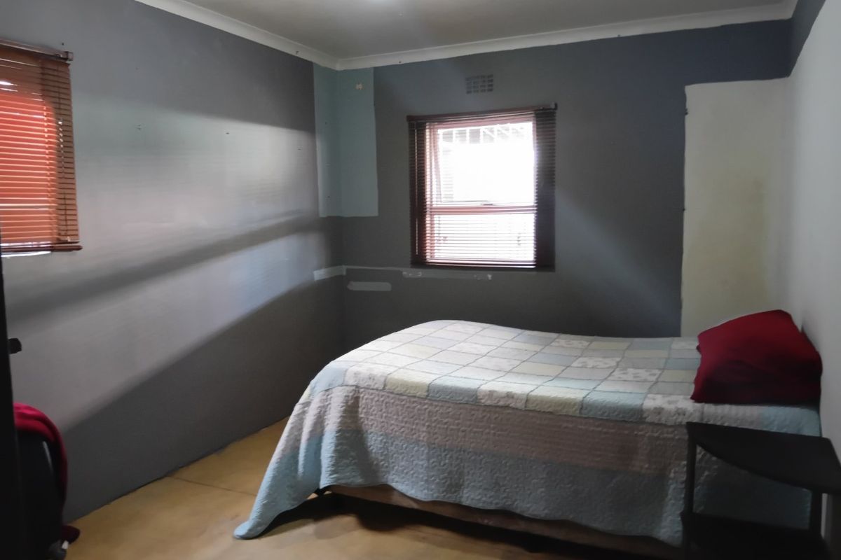 A bedroom featuring grey painted walls with visible patches and unfinished areas, fitted with venetian blinds and a ventilation grille.