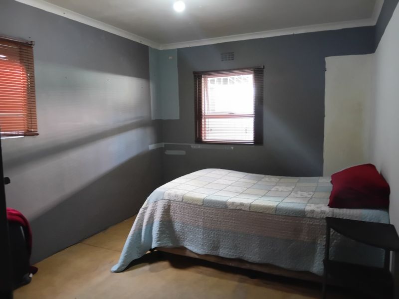 A bedroom featuring grey painted walls with visible patches and unfinished areas, fitted with venetian blinds and a ventilation grille.