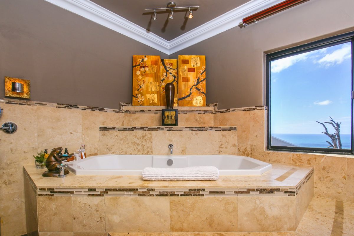 A luxurious bathroom featuring a built-in corner bathtub with travertine tiling, mosaic accents, and a large window offering scenic ocean views.