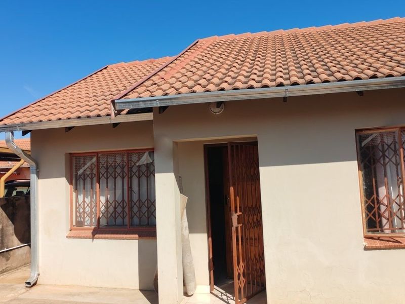 Front elevation of a single-story house featuring a tiled roof, paved driveway, and secure metal gates on the entrance and windows.
