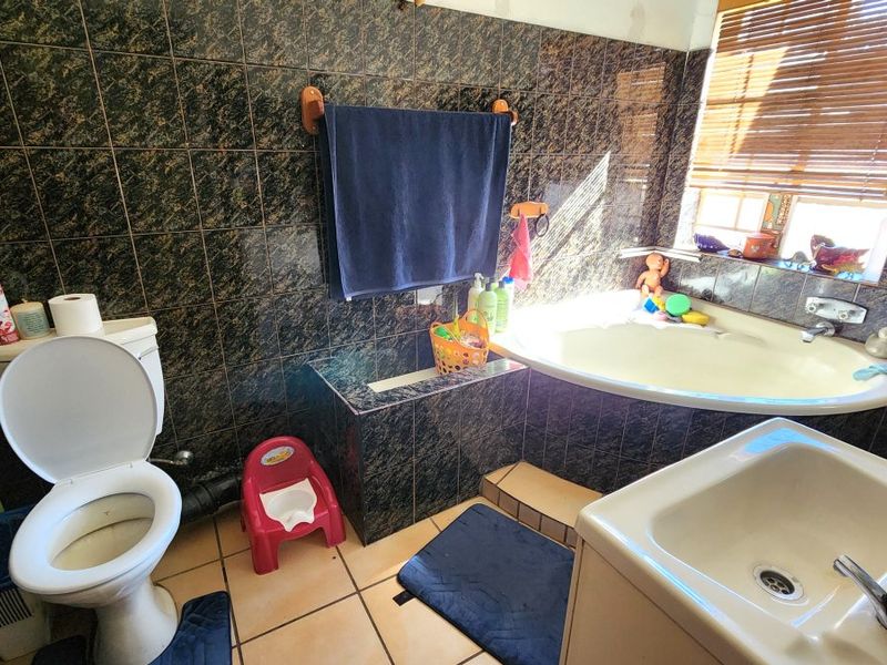 A dated bathroom featuring dark patterned wall tiles, a built-in corner bathtub, a pedestal basin, and a standard toilet.