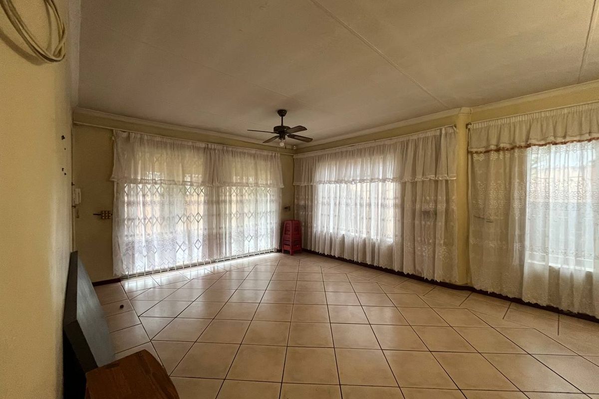 Spacious, empty living area featuring ceramic floor tiles, a ceiling fan, and large windows with security gates, offering a blank canvas for renovation.