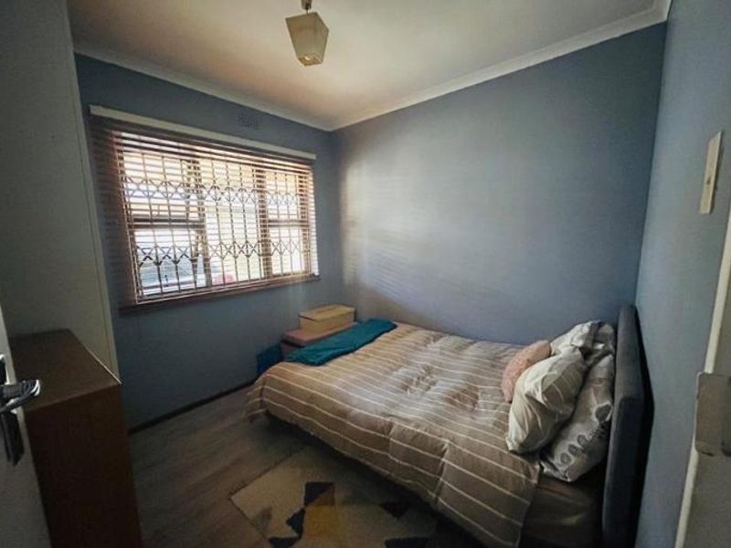 A compact bedroom featuring laminate flooring, blue painted walls, and a window fitted with security bars and venetian blinds.