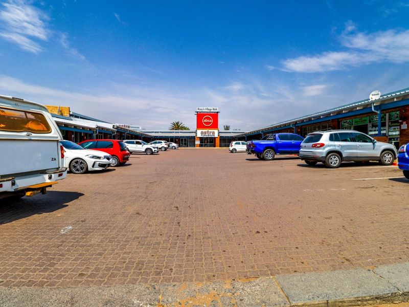 A commercial shopping center exterior featuring a central paved parking lot surrounded by retail storefronts with corrugated metal roofing.