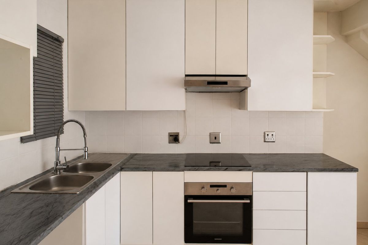 A clean, functional kitchen featuring white cabinetry, dark laminate countertops, built-in oven, electric hob, and a stainless steel double sink with a modern faucet.