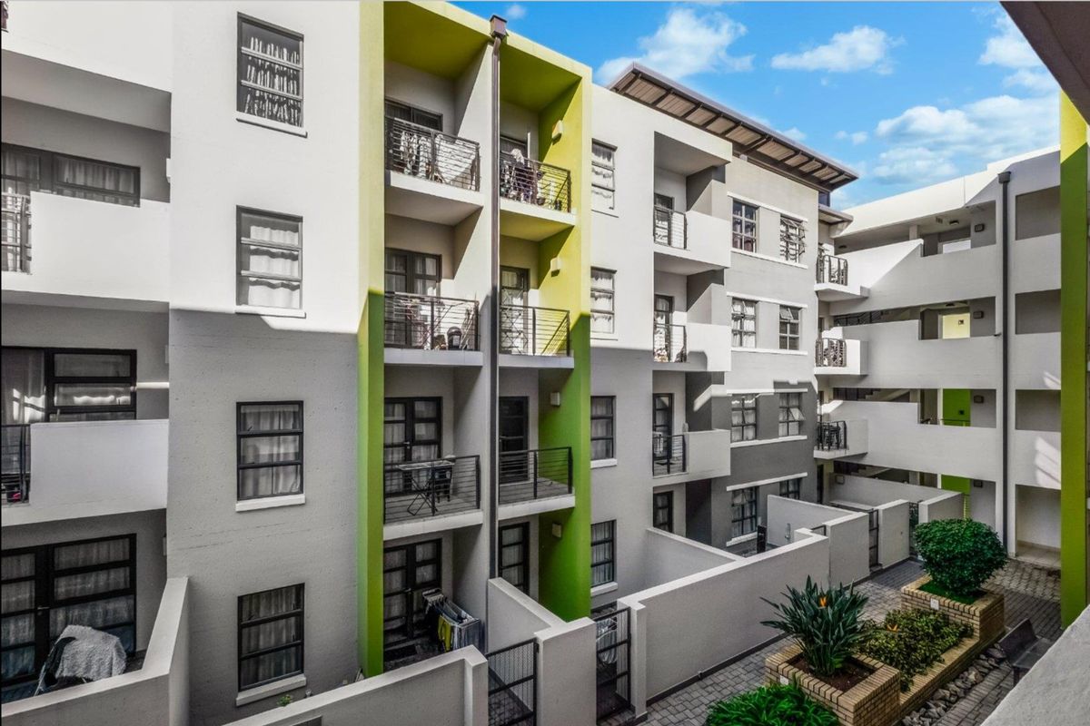 Exterior view of a modern apartment complex featuring stacked balconies with metal railings and a paved ground-level courtyard with brick planter boxes.