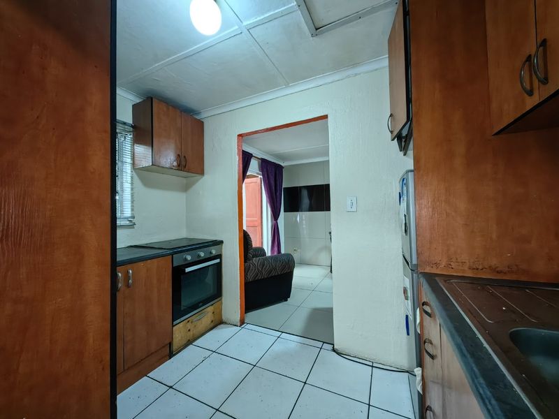 A compact kitchen featuring wooden cabinetry, built-in oven and hob, and tiled flooring, leading through to an adjacent living area.