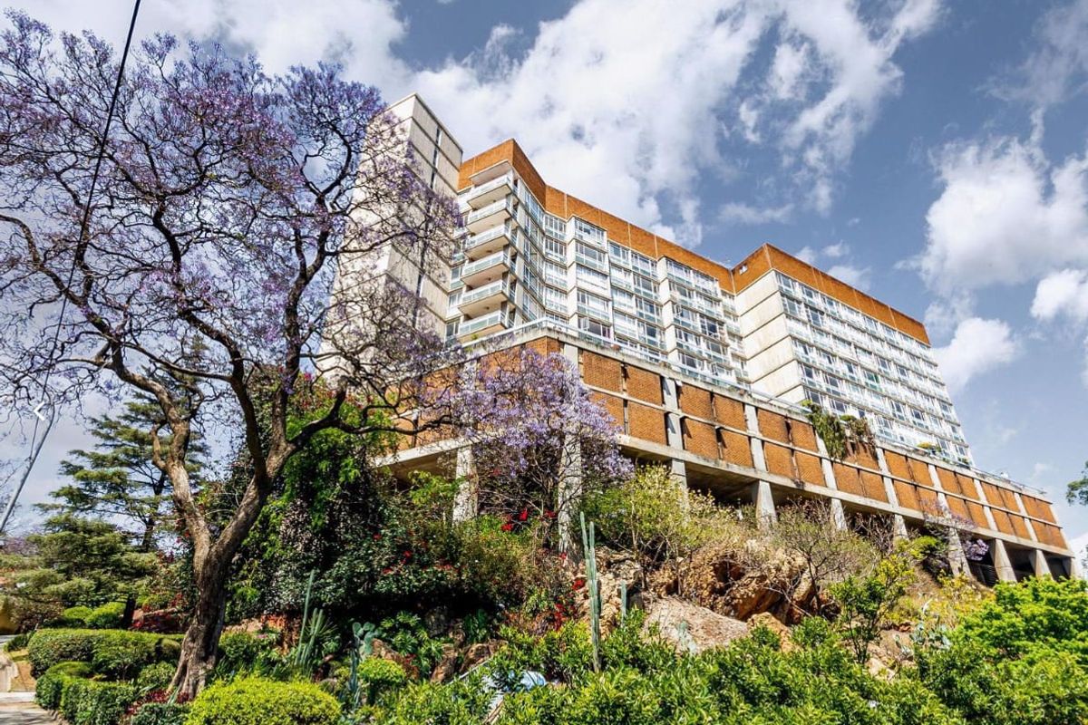 Low-angle view of a multi-story apartment building featuring tiered balconies and a prominent brick and concrete facade set against a landscaped hillside.