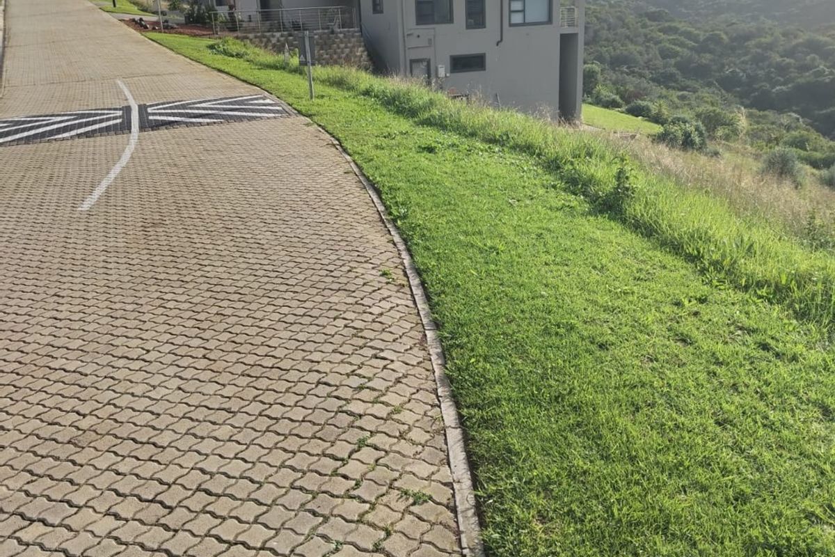 A view of the paved street approach and verge leading towards a residential property set against a hillside landscape.
