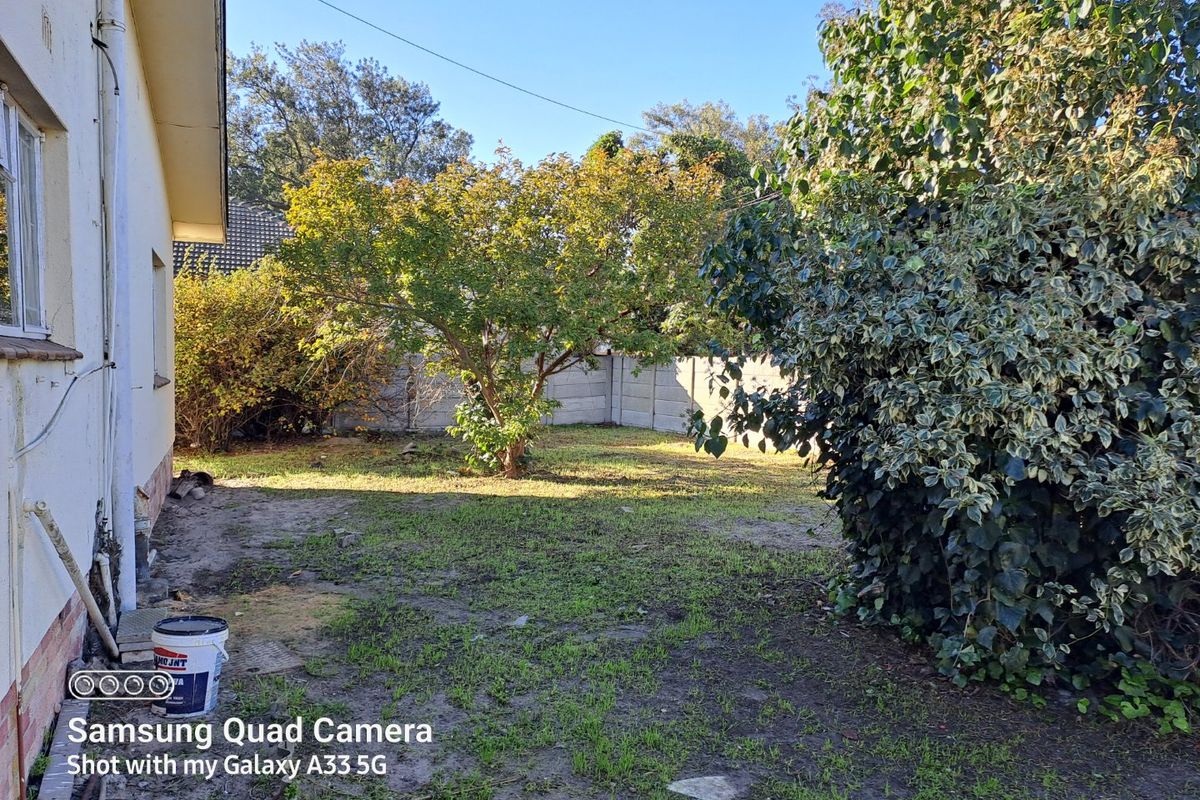 A rear garden area featuring a precast concrete boundary wall, mature trees, and patchy lawn requiring landscaping and maintenance.