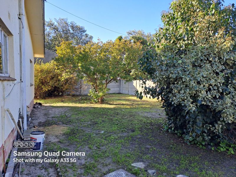A rear garden area featuring a precast concrete boundary wall, mature trees, and patchy lawn requiring landscaping and maintenance.