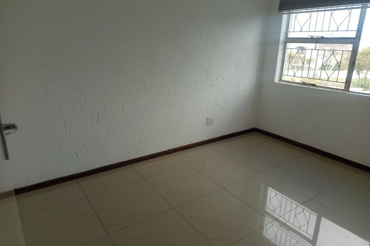 An empty bedroom featuring neutral tiled flooring, textured walls, and a window fitted with security bars and a roller blind.