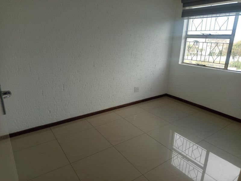An empty bedroom featuring neutral tiled flooring, textured walls, and a window fitted with security bars and a roller blind.