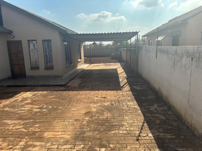 Rear exterior view featuring a paved driveway, a metal carport structure, and a perimeter boundary wall with electric fencing.