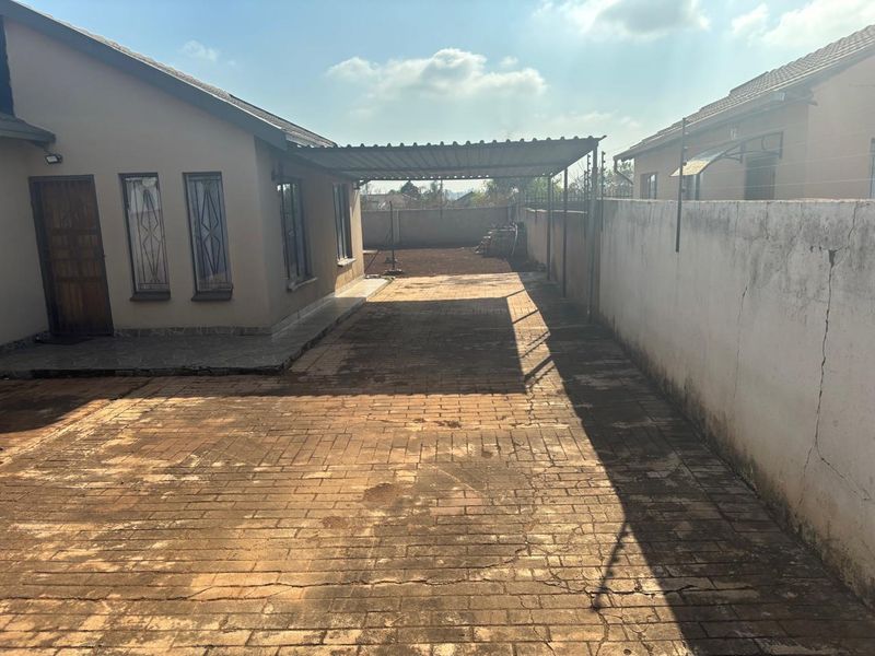 Rear exterior area featuring a paved driveway, a covered carport structure, and a perimeter wall with security features.