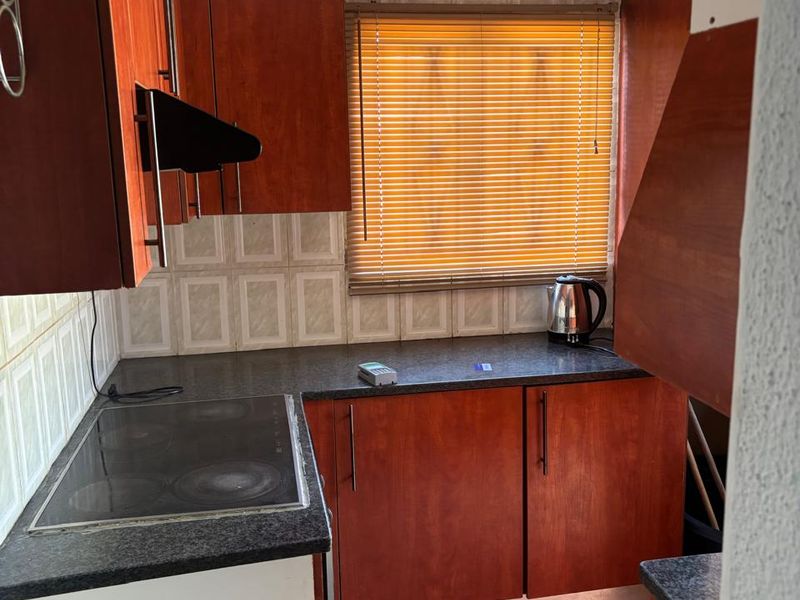 A compact kitchen featuring wood-look cabinetry, a built-in electric hob, tiled backsplash, and tiled flooring, offering a functional layout with potential for modernization.
