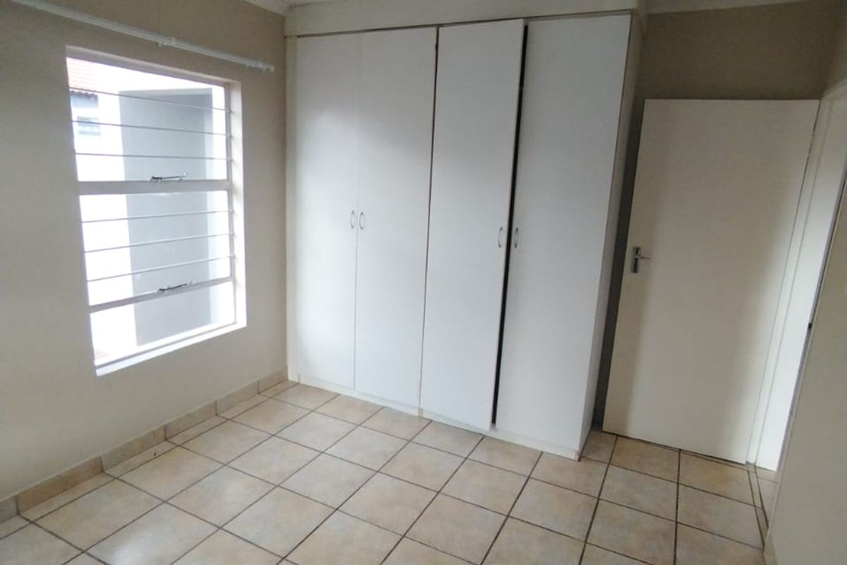 A clean, empty bedroom featuring neutral tiled flooring, built-in white cupboards, and a window fitted with security bars.