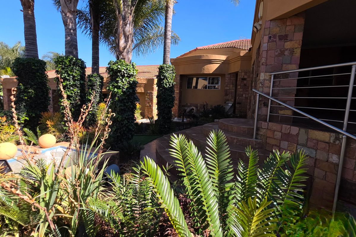 A well-maintained rear exterior view featuring stone-clad walls, a stainless steel balustrade, tiled steps, and established garden landscaping under a clear sky.