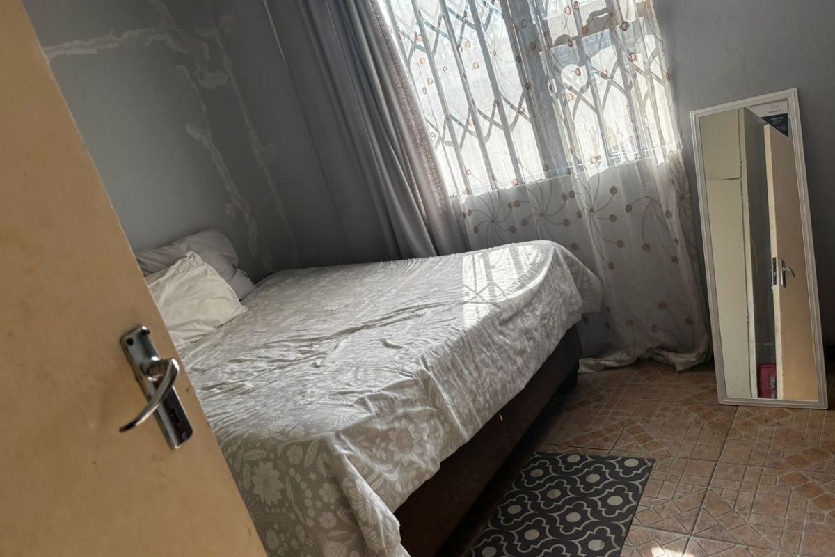 A bedroom featuring tiled flooring and a security gate on the window, currently showing significant wall surface damage and peeling paint.