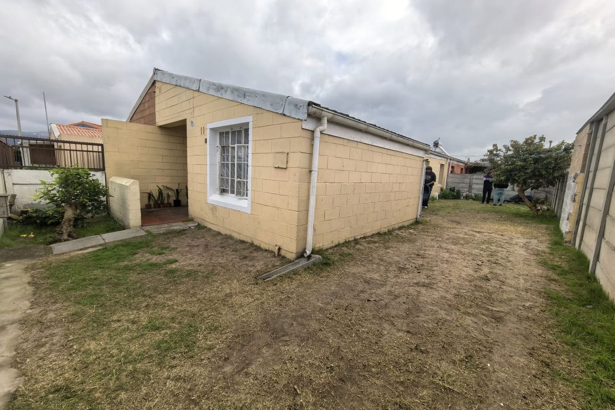 Rear view of a single-story concrete block house featuring a small paved entrance area and a large, unlandscaped backyard with boundary walling.