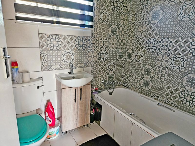A functional bathroom featuring a bathtub, vanity basin, and toilet, accented with decorative patterned wall tiling and a modern roller blind.