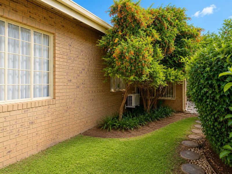 A well-maintained rear exterior view featuring a brick facade, established garden landscaping, stone pathway, and an integrated air conditioning unit.