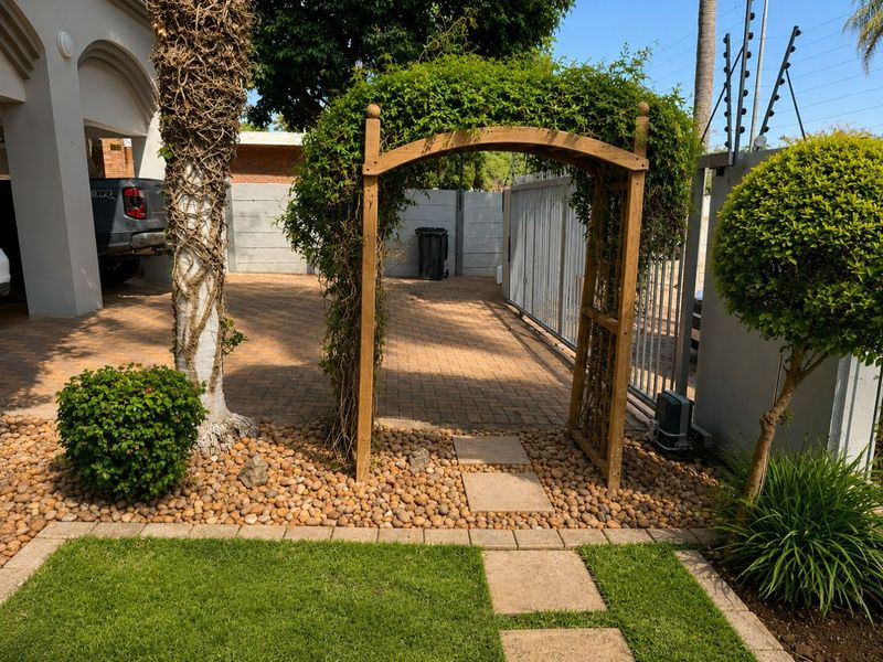 Front entrance area featuring a paved driveway, metal security gate, electric fencing, and a landscaped garden with a wooden archway.