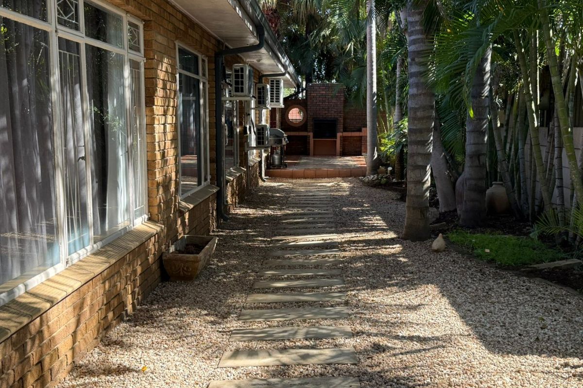 A side garden pathway featuring a brick facade, stone pavers, and a built-in outdoor braai area surrounded by mature palm trees.