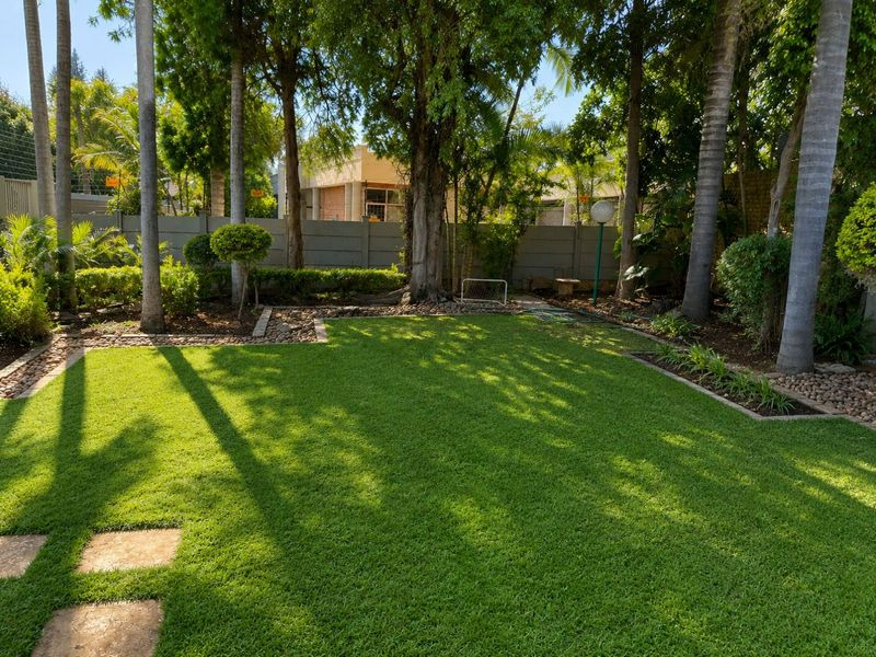 A well-maintained private garden featuring a lush green lawn, mature trees, stone-edged garden beds, and a secure precast concrete boundary wall.