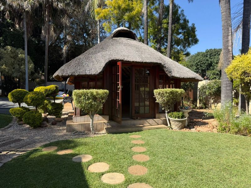 A charming garden setting featuring a traditional thatched-roof structure with timber-framed doors, set within a well-maintained lawn and landscaped outdoor area.
