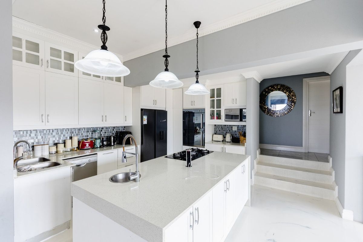 Modern kitchen featuring a central island with quartz countertops, built-in appliances, glass-fronted cabinetry, and elegant crown molding, leading to a raised hallway area.