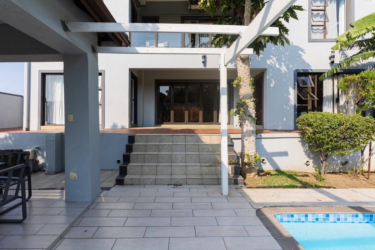 Rear exterior view featuring a tiled patio, swimming pool, and a pergola leading to the main house entrance with sliding glass doors.
