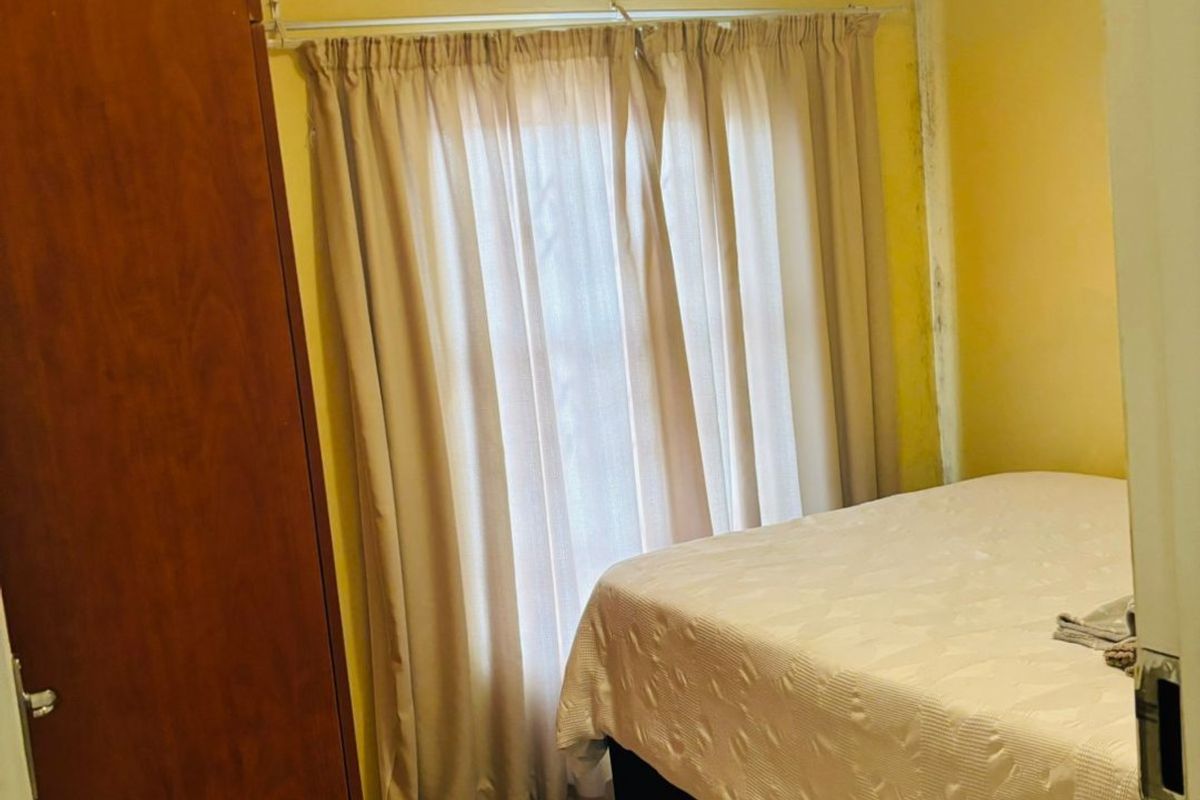 A compact bedroom featuring tiled flooring, a built-in cupboard, and a window with curtains. The room requires cosmetic attention to the walls.