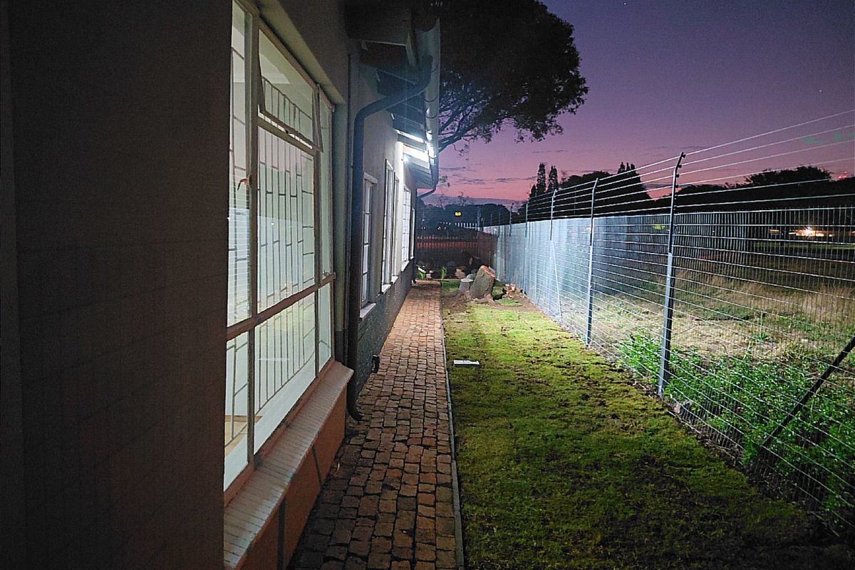 A side exterior view of the property featuring brick paving, security fencing, and external lighting at dusk.