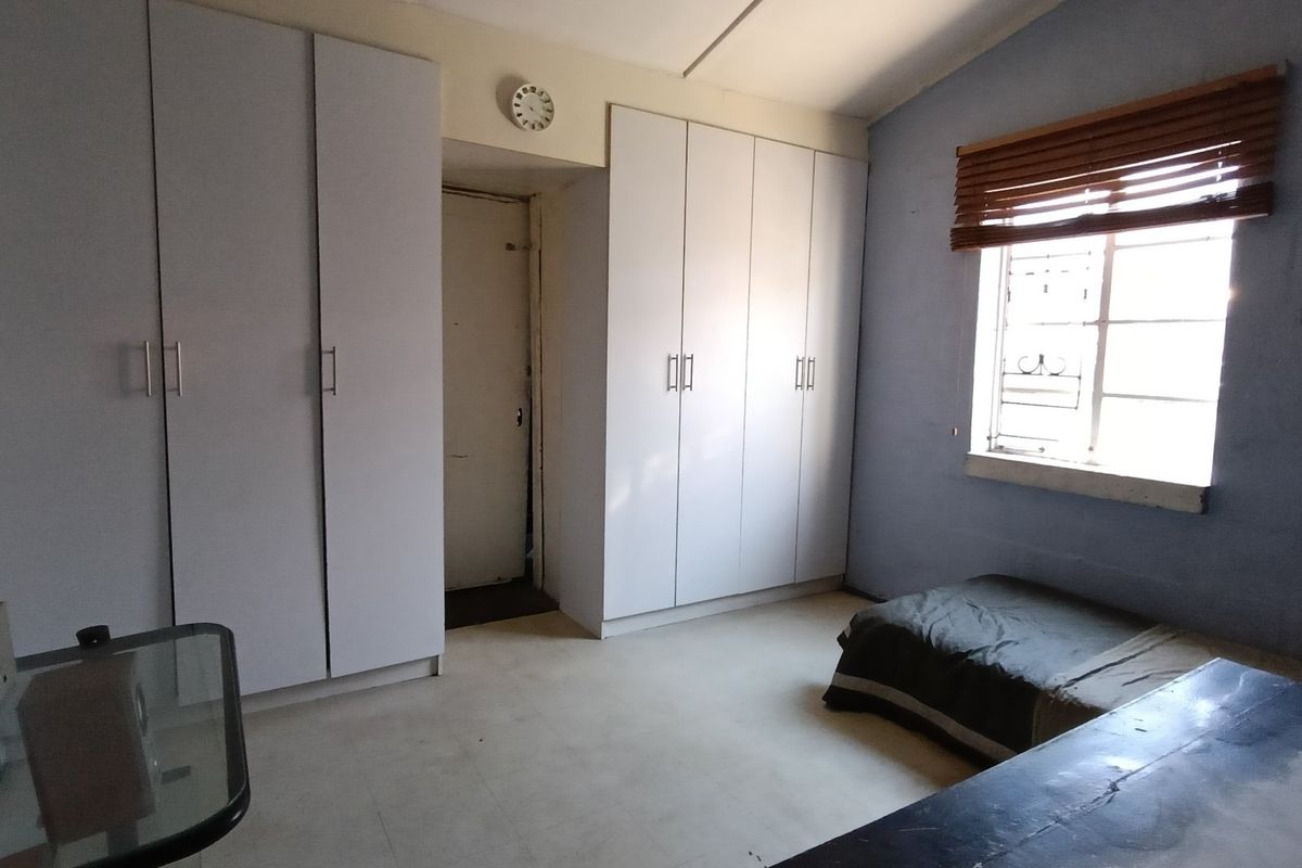A bedroom featuring built-in cupboards, a window with wooden blinds, and security bars, offering a functional space with potential for cosmetic updates.