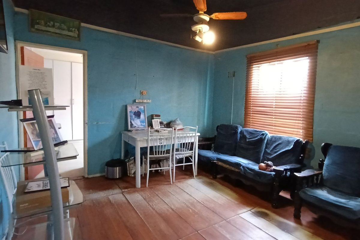 A living room featuring timber flooring, a ceiling fan, and venetian blinds, currently requiring cosmetic updates and wall repairs.