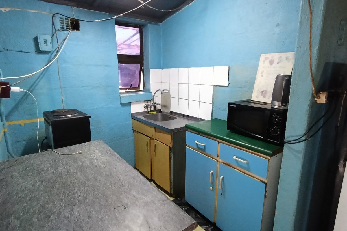 A compact, dated kitchen area featuring basic cabinetry, a stainless steel sink, and tiled splashback, requiring significant cosmetic updates and electrical maintenance.