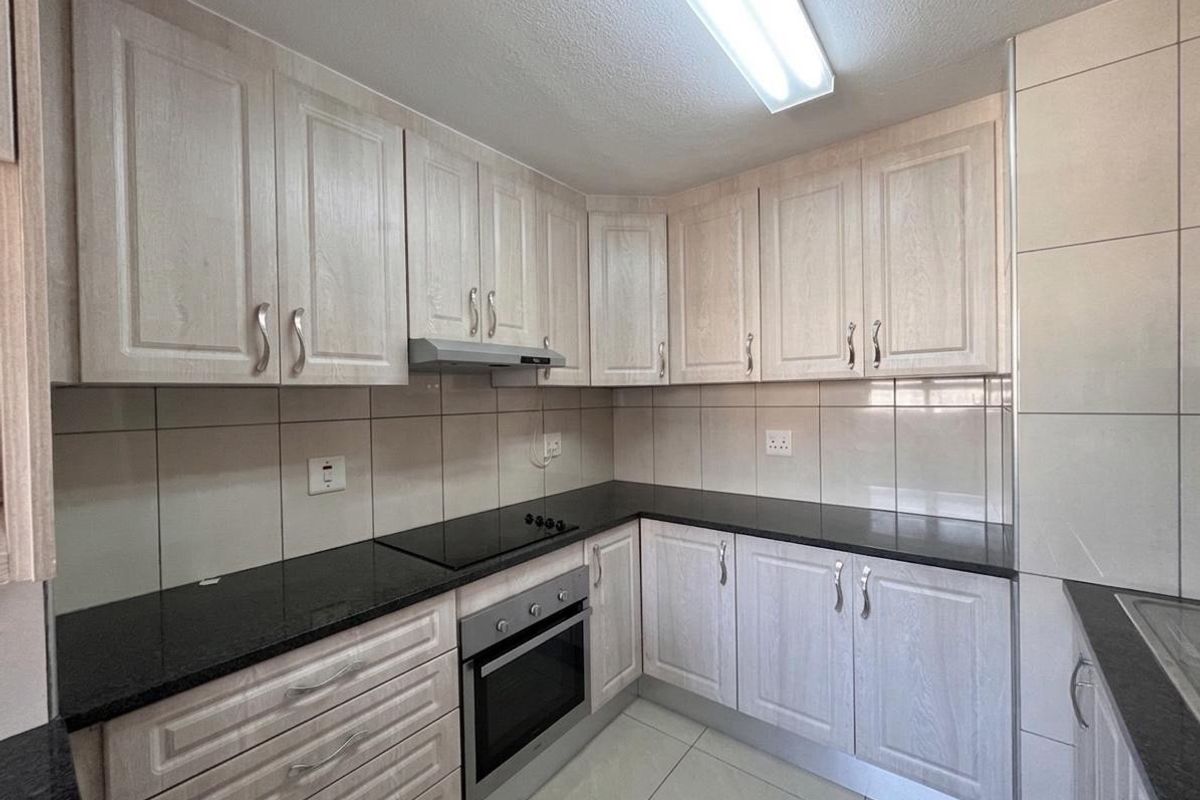 A clean, functional kitchen featuring light wood-finish cabinetry, dark granite countertops, built-in oven, electric hob, and neutral tiled backsplash and flooring.