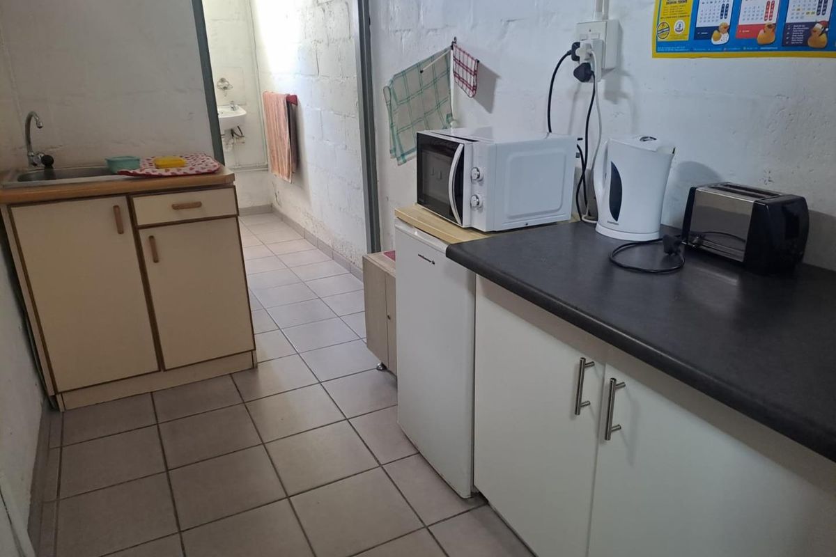 A functional industrial-style kitchenette featuring basic cabinetry, laminate countertops, a stainless steel sink, and tiled flooring with exposed blockwork walls.