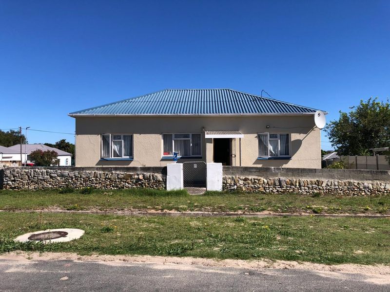 Front elevation of a single-story house featuring a corrugated metal roof, stone boundary wall, and a gated pedestrian entrance.