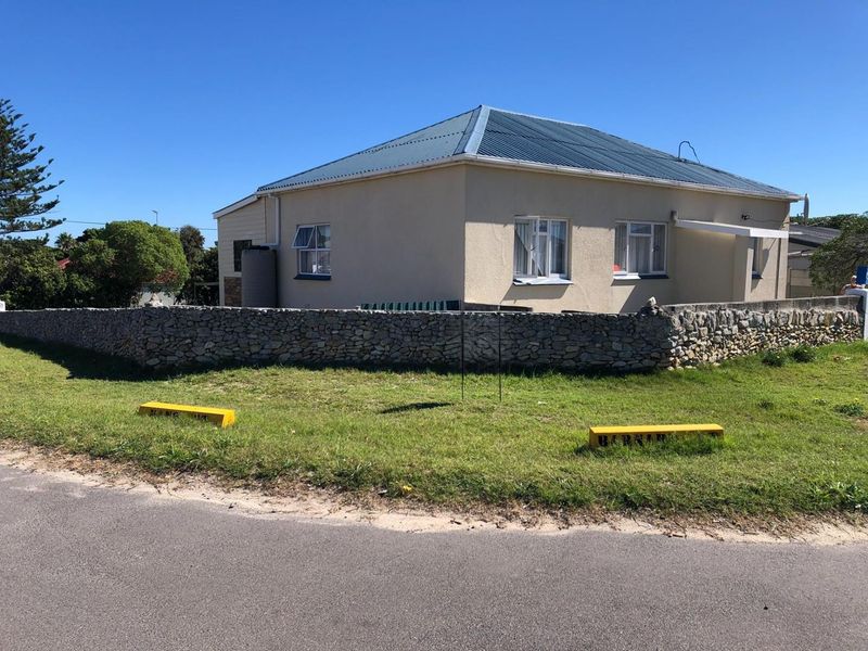 A detached house featuring a stone boundary wall, corrugated metal roof, and aluminium window frames, situated on a corner plot.